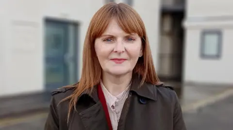 Caoimhe Archibald looks straight ahead at the camera. She wears a brown coat and has red-brown hair and blue eyes. In the background is a white building which is out of focus. 