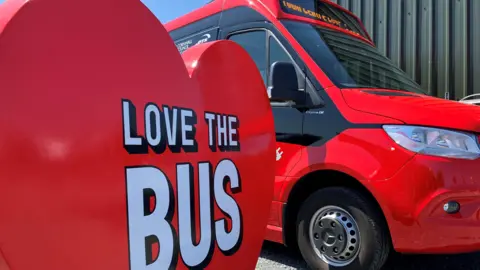 In the foreground is a big love heart promotional stand that says Love the Bus. It is in front of a brand new red electric 18-seater bus.