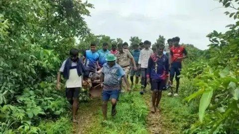 Kayi is carried out of the forest on a stretcher.