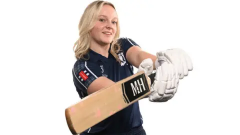 Niamh Muir, who has blonde hair, is wearing a Scotland strip and cricket gloves as she swings a cricket bat