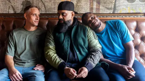 BBC/Tiger Aspect/Khuram Mirza Three characters, Harper (Perry Fitzpatrick), Mobeen (Guz Khan), Nate (Tolu Ogunmefun), sit on a sofa. Two are talking to each other and one has his head on Mr Khan's shoulder, smiling.