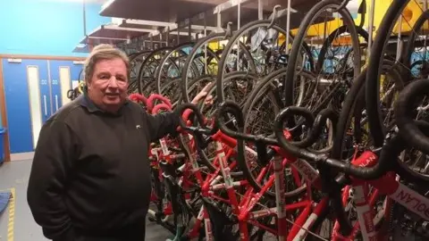 LDRS Alan Davis who has brown hair and is wearing a black jumper, standing next to a rack of red, white and black racing bikes