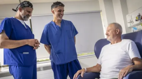 Two men dressed in blue scrubs are on the left and in the middle and standing up. They are looking towards Hall, who has a beard and grey hair and is sitting on a blue chair on the right.