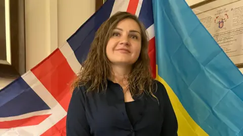 A woman with long brown hair stands in front of a Union Flag and a Ukrainian flag. She is dressed in a navy blue blouse and a black top underneath.