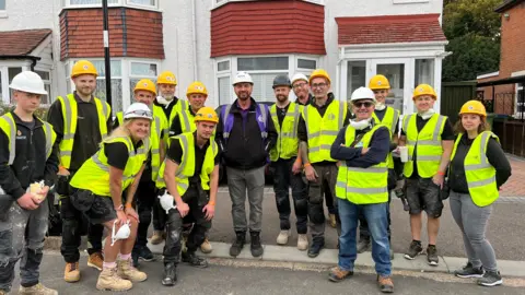 A group of people in yellow florescent yellow vests standing next to TV presenter Nick Knowles outside a home. There's a mixture of men and women all wearing hard hats.