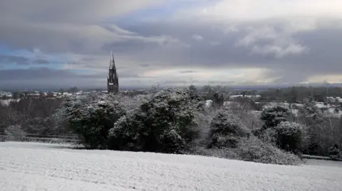 A spire rises above trees in the town of Dungannon with dark grey clouds overhead and a snow-covered field in the foreground 