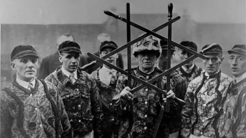 A black and white image of eight sword dancers. They are looking sternly at the camera and one of them is holding six swords in a star formation.
