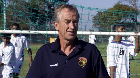 Roald Poulsen pictured in a blue Cape United polo shirt as three young players move a football goal behind him