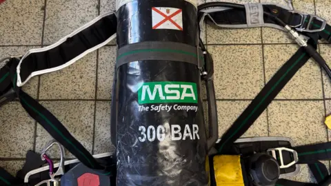 Jersey Fire and Rescue A black compressed air cylinder, part of a self-contained breathing apparatus (SCBA), secured in a harness with shoulder straps and attached components. The cylinder features the MSA logo, "300 BAR" text, and a red and white warning symbol. It rests on a tiled floor with a flashlight and other equipment in the background.