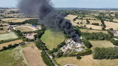 Island Echo A drone view of a fire at a scrapyard surrounded by green fields, with a large plume of black smoke rising above it