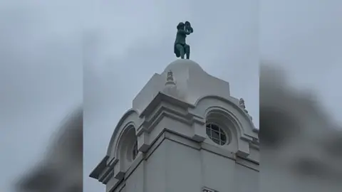 Mark Hall A close up of the small, green dancing lady statue on top of a white domed tower with circular windows. The sky is grey and cloudy.