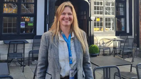 A woman with long blonde hair smiles into the camera. She is wearing a light grey blazer with a white and grey checked shirt and a blue lanyard around her neck. she is standing in front of a medieval-looking building which has modern metal tables and chairs outside it.
