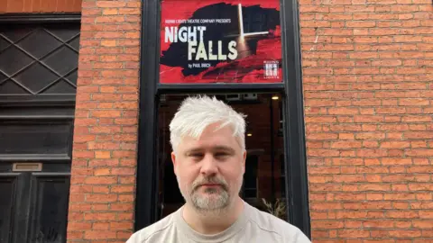 A man with a grey beard standing in front of a brick building. The background features a rectangular poster mounted above a doorway. The poster advertises a theatre production titled “Night Falls”, written by Paul Birch, and includes the name Riding Lights Theatre Company along the top.