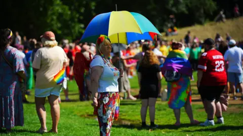 PA Media A shot of lots of people in a park with rainbow gear - focus on a woman holding a large rainbow umbrella to shade herself with a colourful headband too