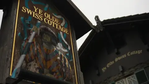 An experienced wood pub sign for BBC "These Olde Swiss Cottage" Characteristic of the depiction of a Swiss guard. In the background, dark shale-style building with gold lettering spelling "These Olde Swiss Cottage",