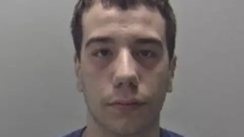 Mugshot of Alexander Jesus, wearing a blue t shirt. He is against a grey backdrop and has short brown hair. He is looking straight into the camera