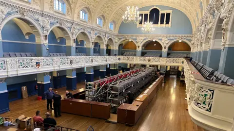 Oxford Town Hall in preparation for the 2024 edition of Oxford Beer and Cider Festival. A few workers are setting up the bar and casks in the middle of the hall. Two chandeliers are lit up on the ceiling.