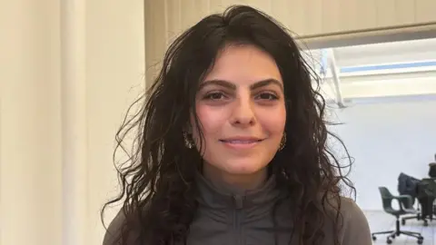 Sarah Dattani Tucker leans forwards and smiles at the camera. She has long curly dark hair worn loose and wears a zip-up grey top. She's pictured in a well lit white-painted room. 