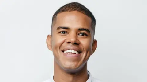 Metropolitan Police Chef Mussie Imnetu in a professional photo. He wears chef's whites and stands against a white background with a broad smile