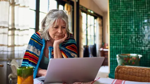 Getty Images An older woman sitting at a table looking at a laptop