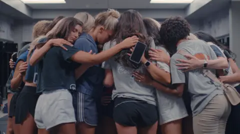 Netflix A large group of cheerleaders looking upset and consoling each other in a huddle