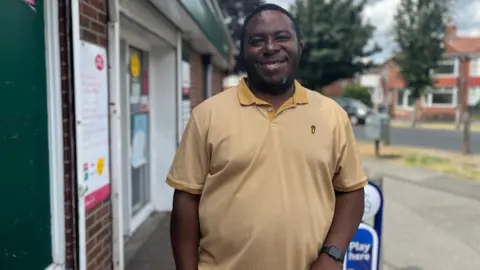 A man with short cropped black hair and beard, wearing a yellow T-shirt. He smiles at the camera in front of the shop.