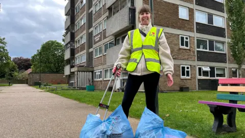 Jack Maclean/BBC Rebecca Fairclough on a path in Mile Cross in Norwich. She is looking directly at the camera and is wearing a cream fleece and black trousers and has a yellow fluorescent vest on over the top. She is holding a little picker and is pointing towards two large blue bags full of litter which has been picked up from the area. In the background is a brightly painted bench and a block of flats.