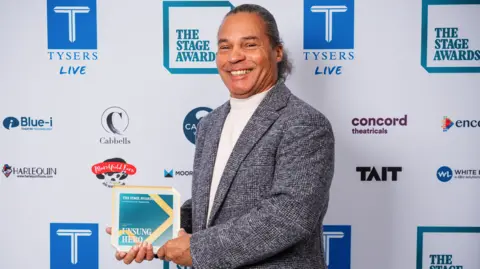 Mike Elliot wearing a white turtleneck jumper underneath a grey tweed blazer. He has long, dark grey hair tied back into a slick low bun. He is smiling and holding his unsung hero award.  The award is a square piece of glass with blue and gold detailing. Behind him is a white banner with the logos of the event sponsors on. 