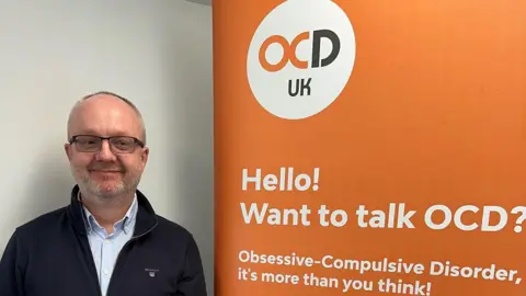 Ashley Fulwood A man with black glasses wearing a blue shirt under a black zip-up stands next to a large orange OCD UK sign. 