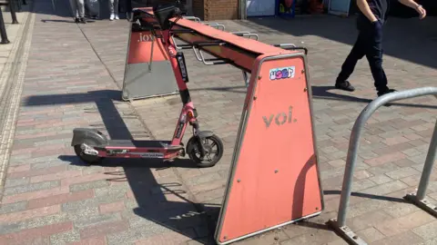 Alex Pope/BBC A Voi e-scooter rack, with a bike on it, in the middle of a town, with people walking past. There are other metal bike racks to the right. 