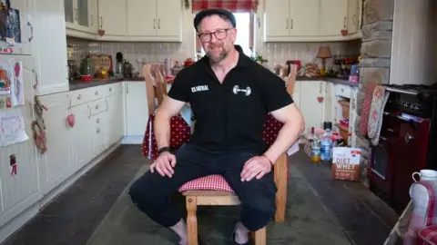 A man in a black polo shirt and flat cap say on a chair in the kitchen. There is a packet of Diet Whey protein behind him