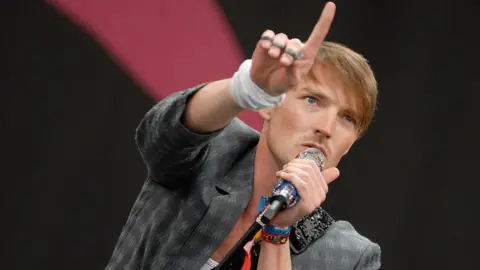 Close up of Dan Gillespie Sells from The Feeling singing on stage. He is holding a mic and pointing with his right hand towards the sky. He has fair hair with a long fringe and a moustache and is wearing a grey blazer
