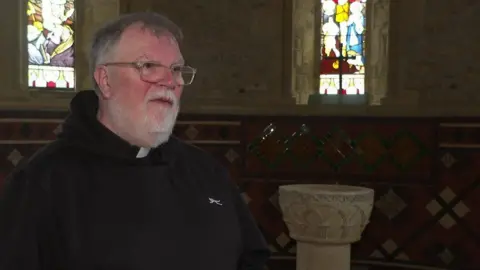 Man with glasses, grey hair and a beard wearing a dog collar and a black top two stained glass windows stand with side of him.