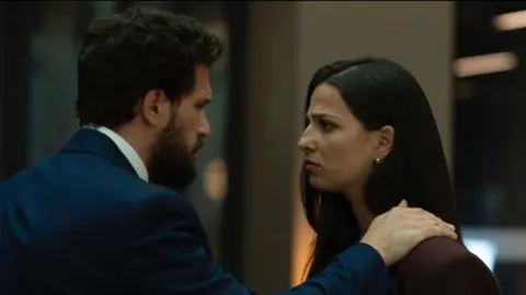 Henry places his hand on Yas's shoulder. They look into each other's eyes