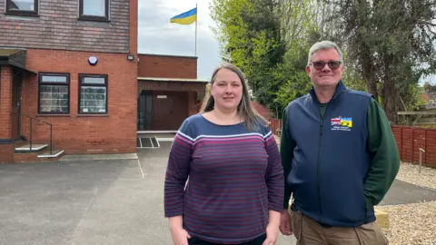 Two people stand in the foreground on the right of the image. A man, William Jackson is wearing a dark blue gilet with a British Ukrainian flag smashing into each other on his chest. He's got grey hair and is wearing sunglasses and tan coloured trousers. Laura Bullivant is next to him she is wearing a striped long sleeved top has brown hair and is a head shorter then him. Behind them are green trees on the right and a building on the left which is a Ukrainian church. Flying atop the church is a Ukrainian flag. They are standing in the churches car park.