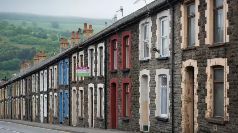 Getty Images A row of terraced homes on a sloped street. They all have stone facades, but have different coloured window sills and porches. 