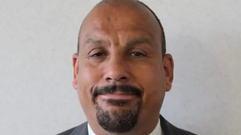 A bald man with a black beard smiling at the camera. He is stood in front of a grey background`