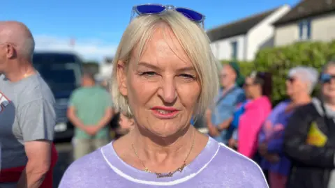 A woman is wearing a purple jumper with white trim. She has sunglasses on the top of her head. She is wearing a necklace and has blonde hair. 