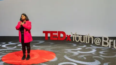 TedX Talks A woman wearing a long pink blazer, black leggings and boots stands on a red circle. The words Tedx Youth Brum can be seen behind her.