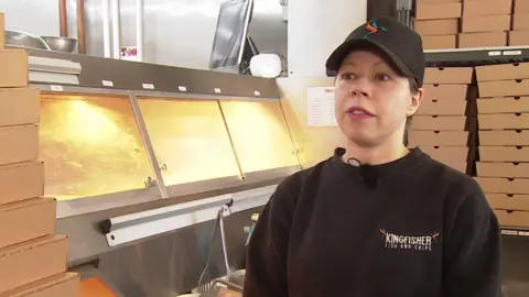 Liza Salazar is standing in her fish and chip shop in front of the food display. She is wearing a black cap and black sweatshirt and has her brown hair tied back. There are takeaway boxes piled up behind her.