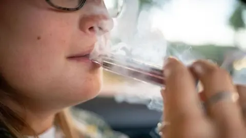 A young woman exhales a cloud of vapour while placing a vapouriser to her lips while she sits in a car. Bright light comes in through the window behind her.
