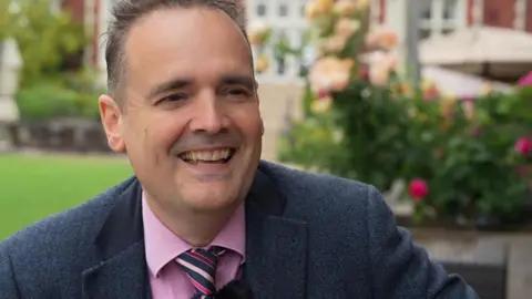 A man smiling widely and looking past the camera to the right. He is wearing a jacket, shirt and tie. There is a garden in the background.