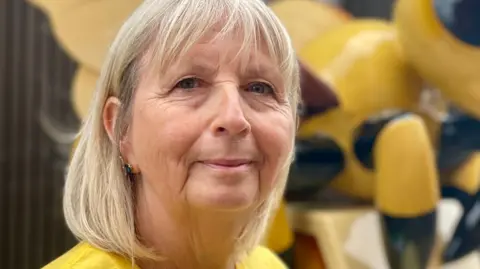 Steve Huntley/BBC Pauline Harvey-Jones has a blonde bob, and is wearing a yellow T-shirt. Part of a yellow and black bee sculpture can be seen behind her.