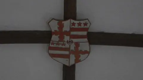 A white and red shield attached to the ceiling split into four sections. Top right and bottom left is three red stars above two red stripes which is the Washington family's coat of arms.