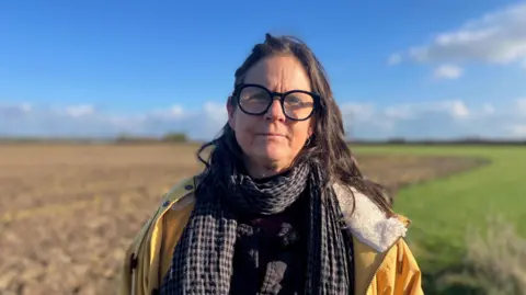 A woman wearing glasses looks at the camera. She is wearing a brown and black chequered scarf and a yellow anorak. Behind her is a brown ploughed field and a strip of grass.