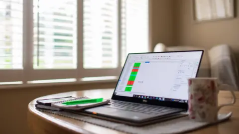 An open laptop on a table with a mug, pen and notebook placed next to it. 