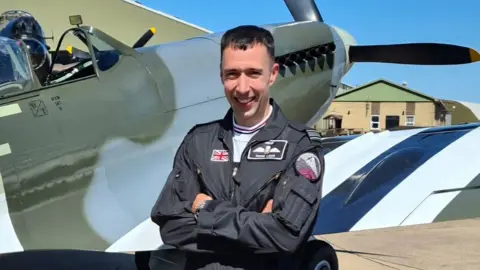 RAF / Crown Copyright Sqn Ldr Mark Long in dark blue RAF flying suit standing with arms folded in front of a Spitfire aircraft