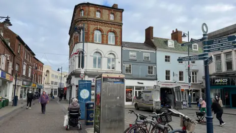 A photograph of Boston town centre.  A telephone box is in the middle of the picture.  Bikes are leaning up against a bike stand and a pensioner is moving along the paved area in a mobility chair.  