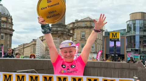 Alice Townsend smiling into the camera with her arms up in the air. She is wearing a colourful cap with the brim standing up and has white hair peeking through. She is wearing a bright pink t-shirt and is holding a large cardboard medal in one of her hands which reads "Alice Townsend Metro Marathon". People are holding a white finishing line banner in front of her with the yellow Metro logo, which is a black capital M in a yellow box. The bottom of the stone Grey's Monument is behind her, as well as a large Metro sign. The stone buildings of Grainger Street are also behind her along with the glass-fronted Eldon Square. 