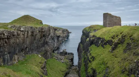 Stacked grey and brown rocks sit on top of a green hilltop where the dark blue sea enters a gully. The background is overcast with light grey clouds 